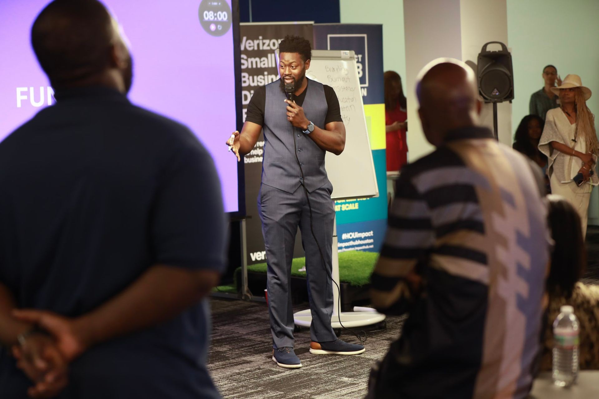 Person speaking into a microphone during a presentation at a business event with an audience in the foreground.