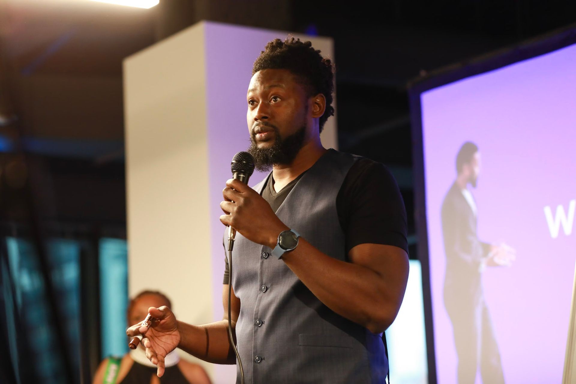 Man in a vest speaking into a microphone on stage with a projected screen in the background.