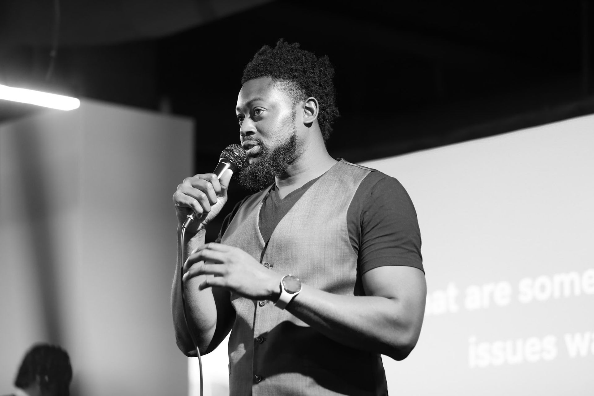 Person wearing a vest and a wristwatch, holding a microphone and speaking on stage.